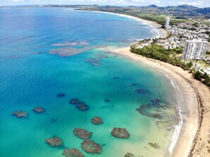 An aerial view of a coastal area with clear turquoise waters, a sandy beach, and a residential area with buildings and greenery.
