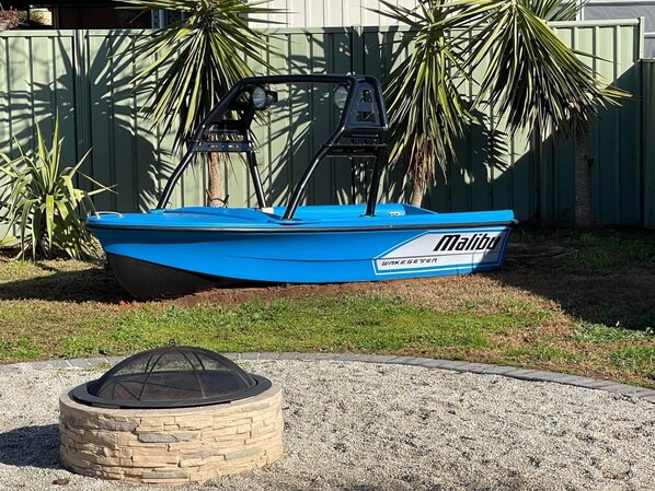 A blue boat with the brand name Malibu and WakeSetter on it, parked in a grassy area with a stone fire pit nearby.
