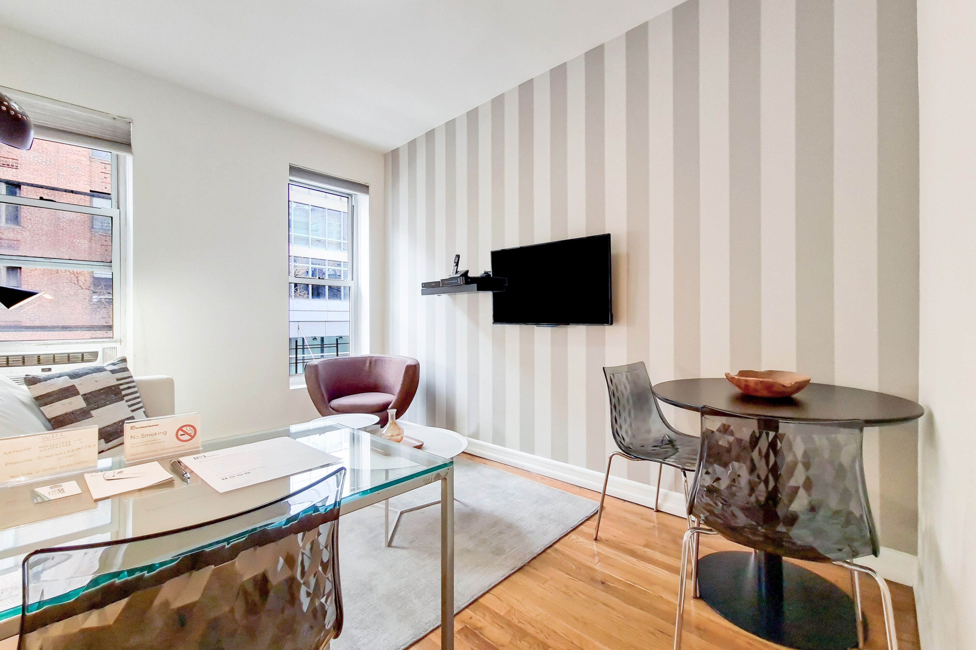 Una sala moderna con una mesa de centro de cristal, un sillón rojo, una mesa redonda con un bol, un televisor montado y una pared a rayas.