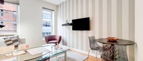 Una sala moderna con una mesa de centro de cristal, un sillón rojo, una mesa redonda con un bol, un televisor montado y una pared a rayas.
