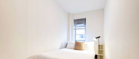 A small, neatly arranged bedroom with a bed, a window with blinds, a bedside table, and a floor lamp.