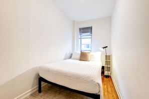 Un dormitorio pequeño y ordenado, con cama, ventana con persianas, mesita de noche y lámpara de pie.