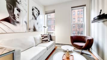 A modern living room with a white sofa, a red armchair, and a coffee table. There are two windows with views of buildings.