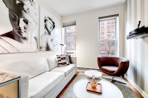 Un salon moderne avec un canapé blanc, un fauteuil rouge et une table basse. Il y a deux fenêtres donnant sur des immeubles.