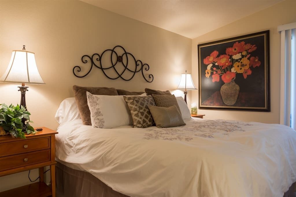 A neatly made bed with a white duvet, multiple pillows, and a decorative wall piece above it. There are two bedside tables with lamps and a potted plant on one.