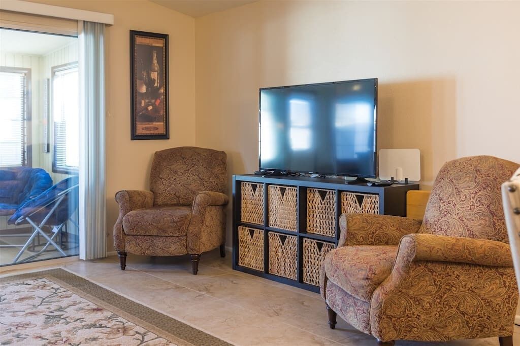 A living room with a large flat-screen TV, patterned armchairs, a glass door leading outside, and a framed picture on the wall.
