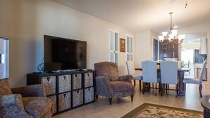 Un salon comprenant un téléviseur, des fauteuils à motifs, un coin repas avec des chaises et un lustre.