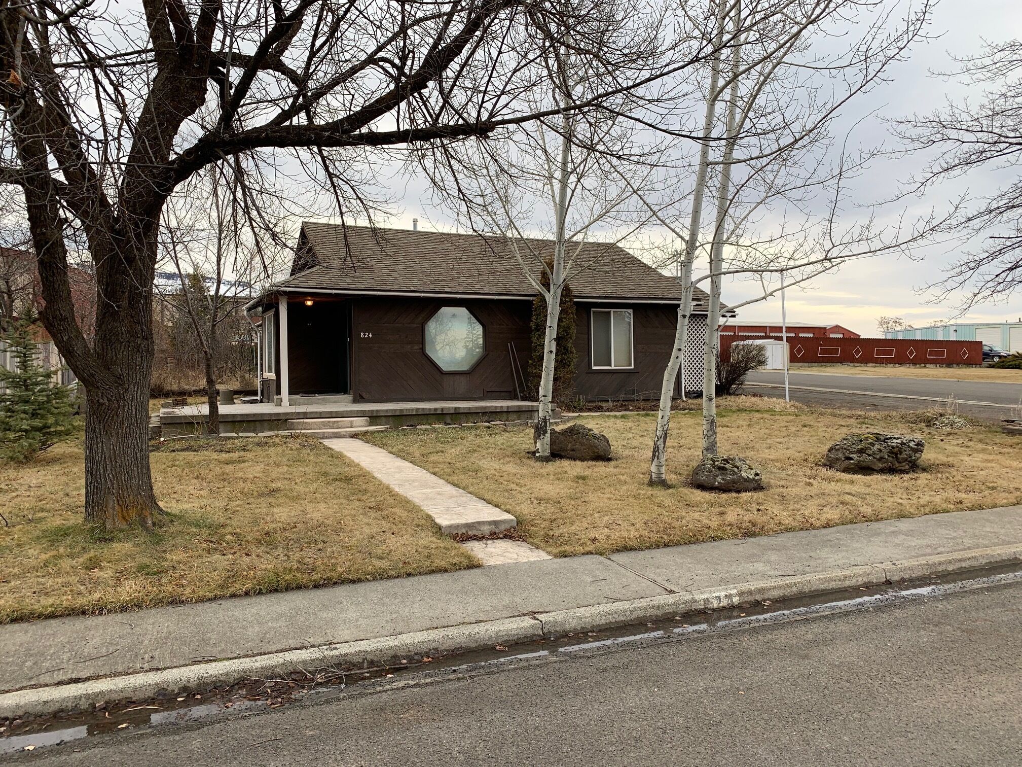 A single-story house with a dark brown exterior, a garage door, and a small front yard with a few trees and rocks.