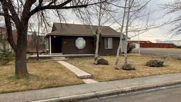 A single-story house with a dark brown exterior, a garage door, and a small front yard with a few trees and rocks.