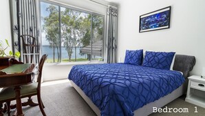 A bedroom with a large bed, a desk, and a chair. There is a window with curtains and a view of trees and a building. A painting is on the wall. - Getaway Lakefront environmental House with Kayaks Water Views (Chain Valley Bay)