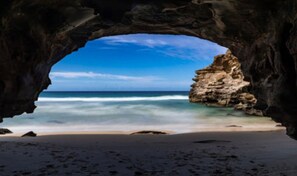 A beach view through a natural rock arch with clear blue water and a sandy shore. - Getaway Lakefront environmental House with Kayaks Water Views (Chain Valley Bay)