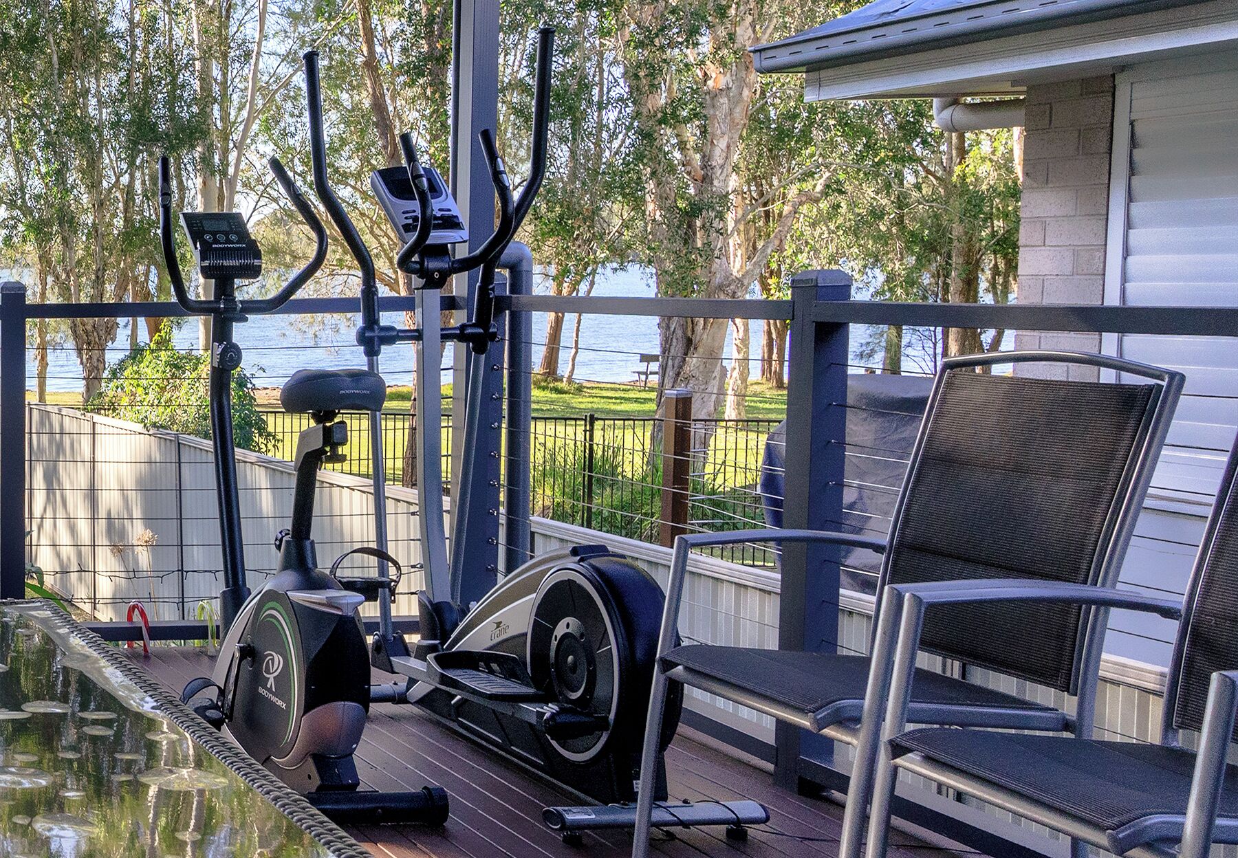 An outdoor exercise area with a stationary bike, elliptical, and rowing machine, a chair, and a view of the water.