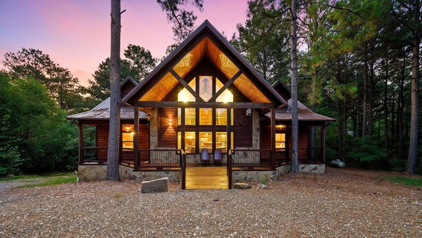 A log cabin with a wooden exterior, a stone foundation, and a wooden deck with chairs. - Spacious 8 BR Broken Bow Family Cabin with Hot Tub, Fire Pit, Arcade, Swing (Broken Bow)