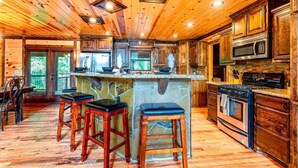A rustic kitchen with wooden cabinets, a central island with stools, a stove, and a microwave. - Spacious 8 BR Broken Bow Family Cabin with Hot Tub, Fire Pit, Arcade, Swing (Broken Bow)