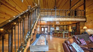 A spacious living area with a foosball table, a brown leather couch, and a staircase with a railing. - Spacious 8 BR Broken Bow Family Cabin with Hot Tub, Fire Pit, Arcade, Swing (Broken Bow)