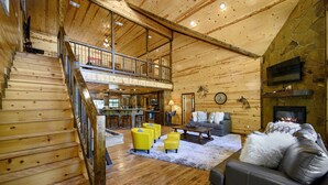 A spacious wooden interior with a staircase leading to a loft, a fireplace, and a cozy seating area. - Spacious 8 BR Broken Bow Family Cabin with Hot Tub, Fire Pit, Arcade, Swing (Broken Bow)