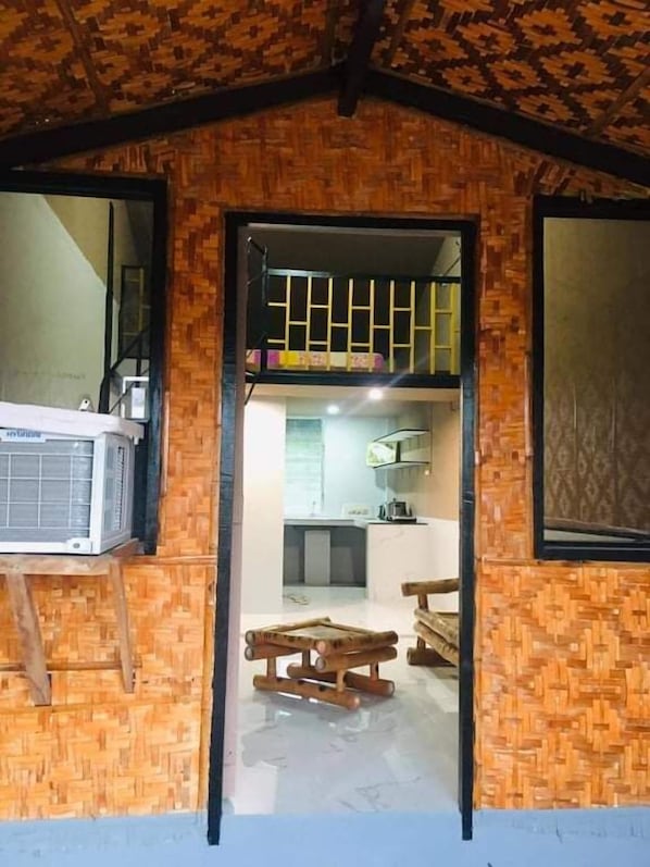 Une pièce avec un plafond haut et un mur orné d’un motif tissé. On y trouve un lit avec un climatiseur, un banc en bois et une table.
