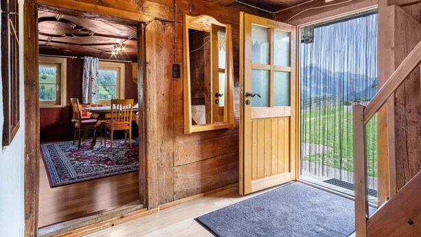 A wooden cabin interior with a dining area, a wooden door leading outside, and a view of the outdoors.