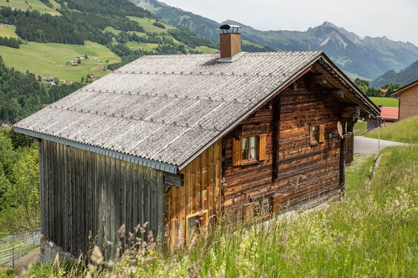 A rustic wooden cabin with a sloped roof, situated in a mountainous area with a view of green valleys and distant peaks.