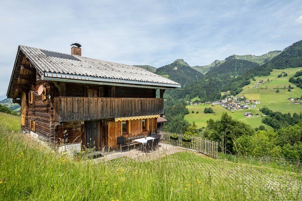 A wooden cabin with a balcony, set on a hillside overlooking a valley with houses and mountains.