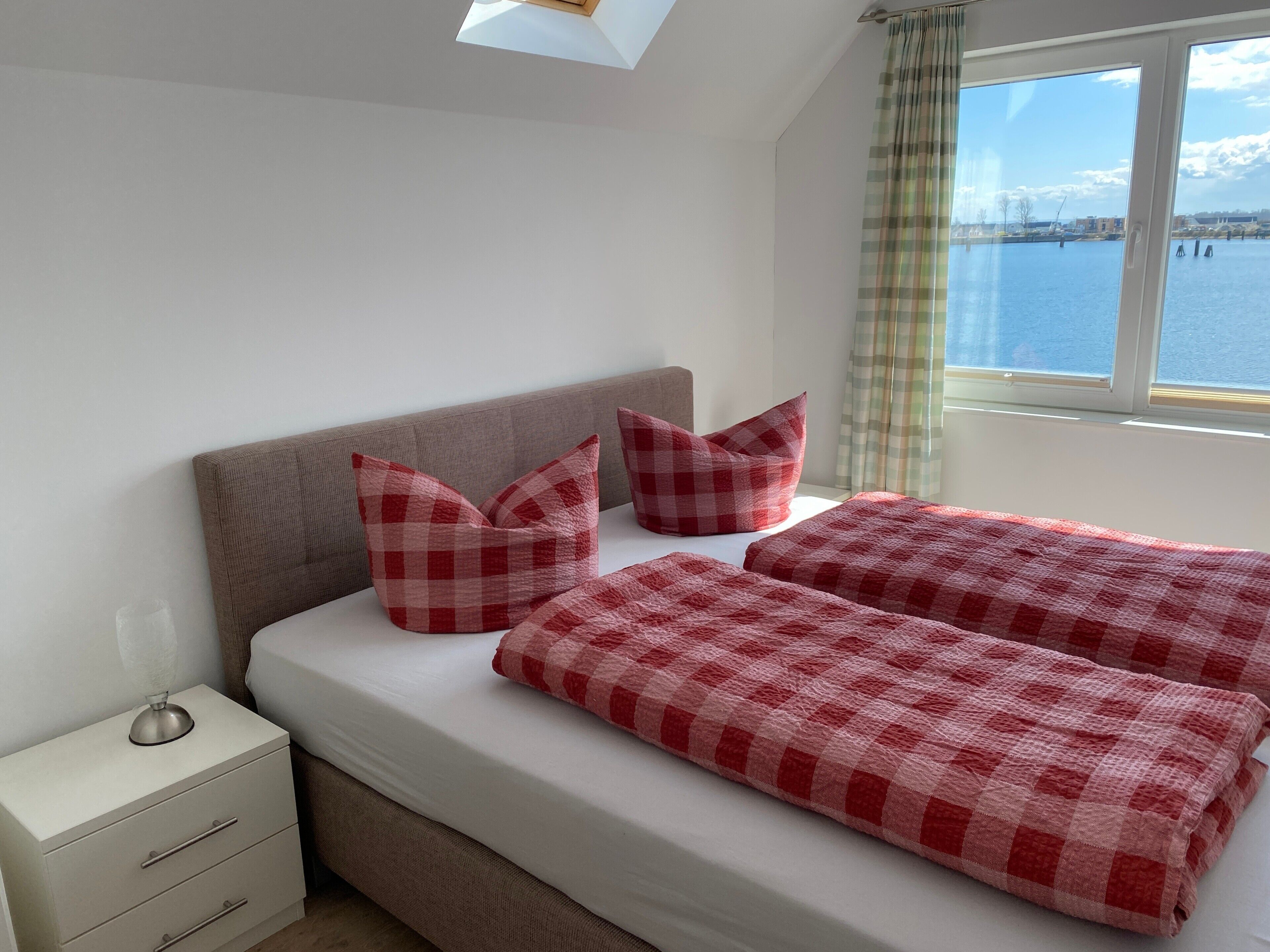 A bedroom with a bed, pillows, a nightstand, and a window with a view of a body of water.