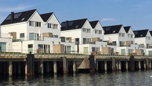 Une rangée de maisons modernes aux façades blanches et aux toits noirs, reliées par une jetée qui surplombe l’eau.
