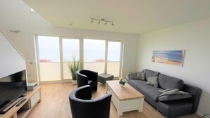 Un salon moderne doté d’un téléviseur à écran plat, d’un canapé, d’une table basse et d’une grande fenêtre offrant une vue sur la mer.