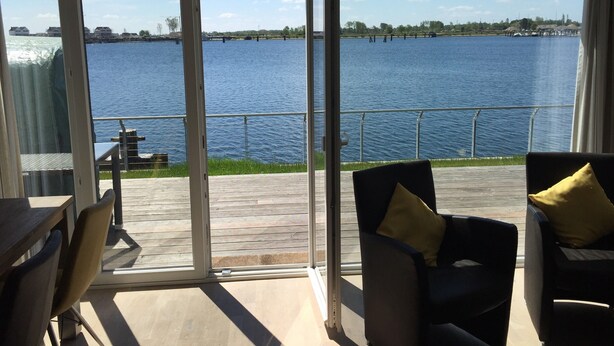Een kamer met uitzicht op een meer, een eethoek en een zithoek met stoelen en kussens.