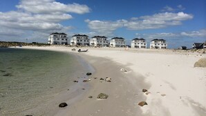 Un hôtel situé en bord de mer, composé de plusieurs bâtiments blancs, avec une plage de sable fin et un ciel d’un bleu éclatant.