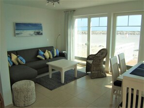 A living room with a sofa, a coffee table, a wicker chair, and a view of the beach.