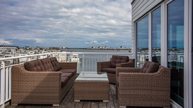 A balcony with wicker furniture, a glass table, and a view of the city and water. - Vacation Home "Lootsen Hus by Seeblick Ferien" with Sea View (OstseeResort Olpenitz/ Kappeln)