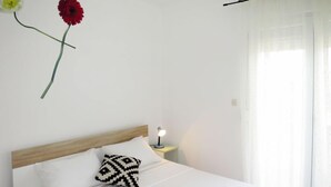 Een net opgemaakt bed met witte lakens en een kussen met een patroon, een bedlampje, en een enkele rode bloem op een stengel aan de muur.
