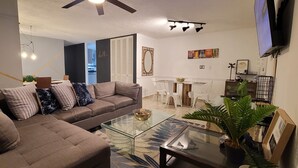 Una sala de estar con un sofá modular, una mesa de centro de vidrio, un comedor con una mesita y sillas, y un televisor montado en la pared.