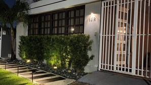 Un edificio moderno con entrada protegida por un portón, un seto cuidado y una escalera que conduce a la entrada.