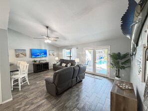 A spacious living room with a gray sofa, a dining area with a white table and chairs, a ceiling fan, and a view of a backyard with a pool.