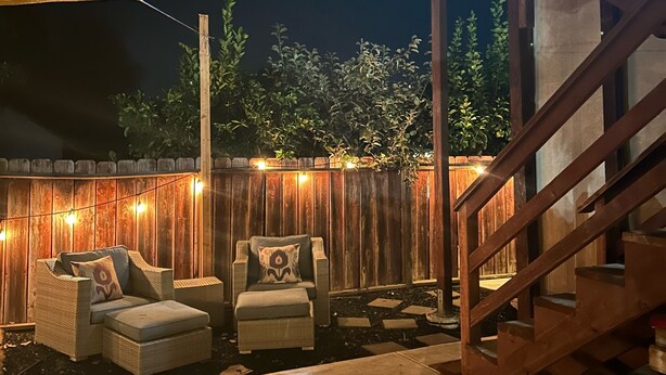 Un patio al aire libre con terraza de madera, dos sillas de mimbre, una mesita y una escalera de madera que lleva a un nivel superior.