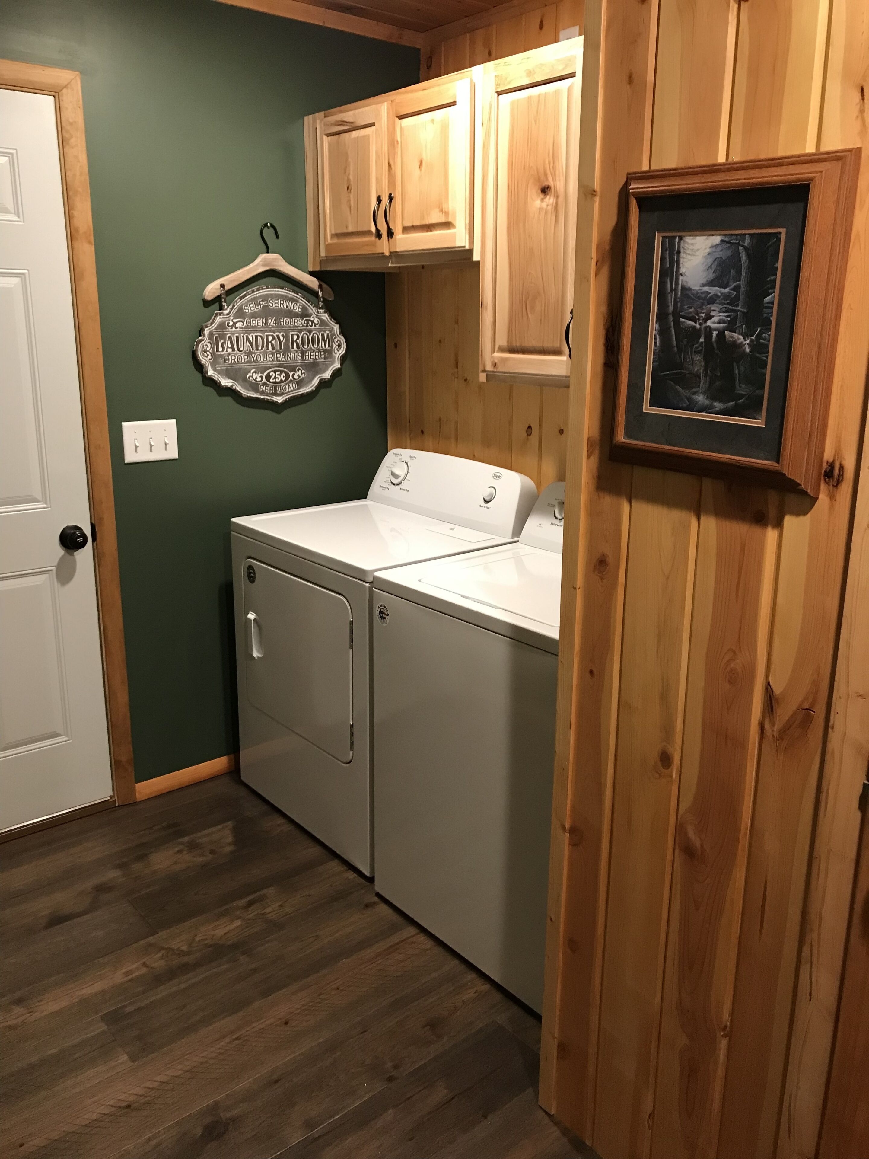 A laundry room with a front-loading washer and dryer, wooden cabinets, a framed picture on the wall, and a sign indicating self-service laundry.