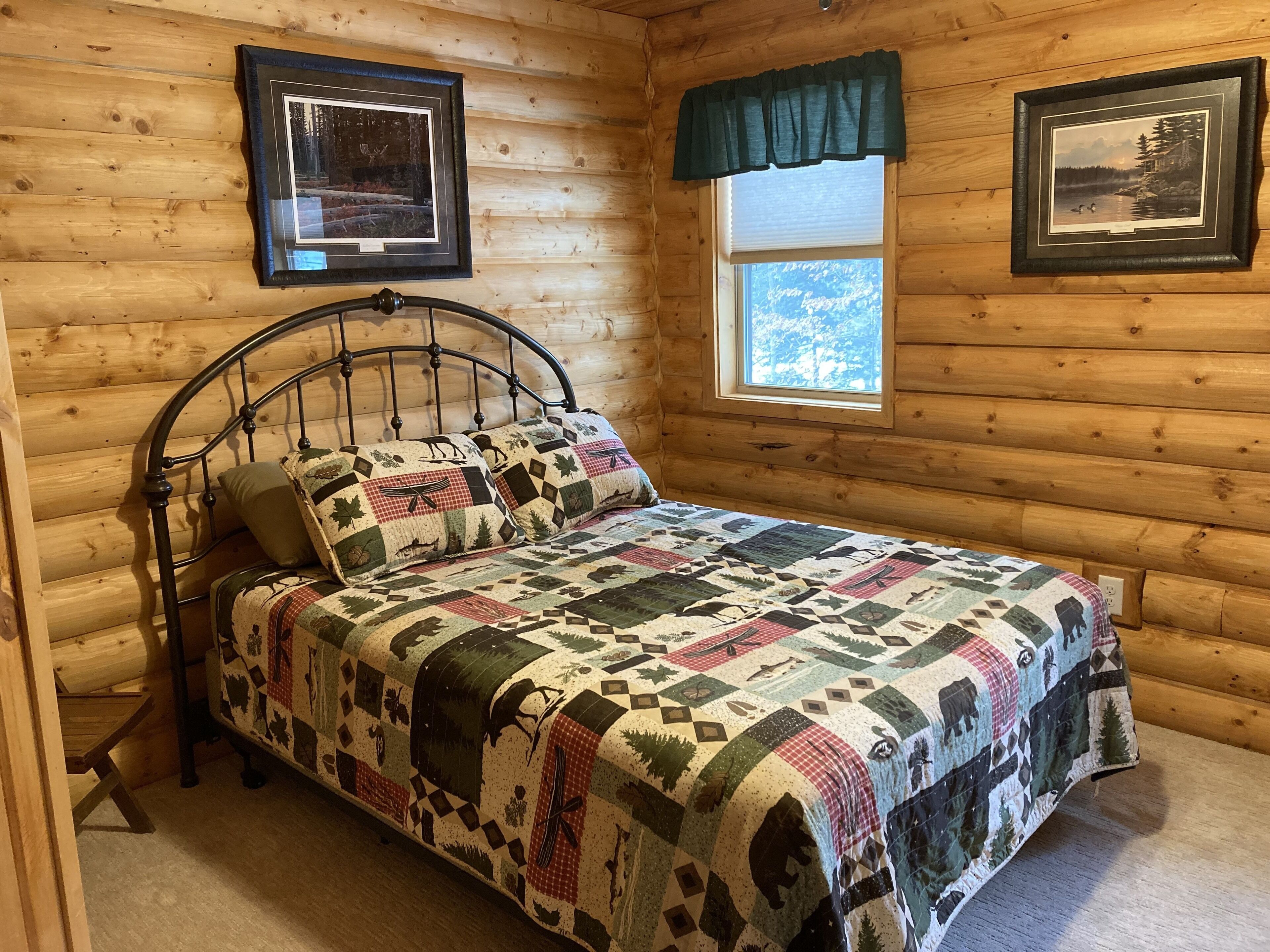 Ein Schlafzimmer in einer Holzhütte mit einem Metallbettgestell, einer Steude mit Wald- und Tierdrucken, zwei gerahmten Bildern an den Wänden und einem Fenster mit Blick auf Bäume.
