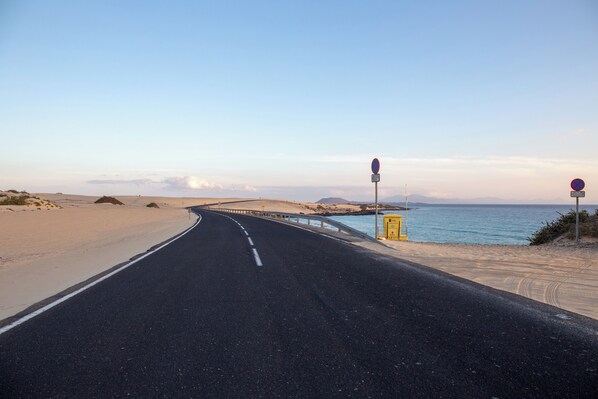 A coastal road with a clear sky, a sandy beach, and a sea view. - Casa Tiago With High-speed Wifi and Smart-tvv (La Oliva)