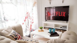 A living room with a flat-screen TV displaying the Netflix logo, a white coffee table with books and a plant, and a sofa with a cushion. - Casa Tiago With High-speed Wifi and Smart-tvv (La Oliva)