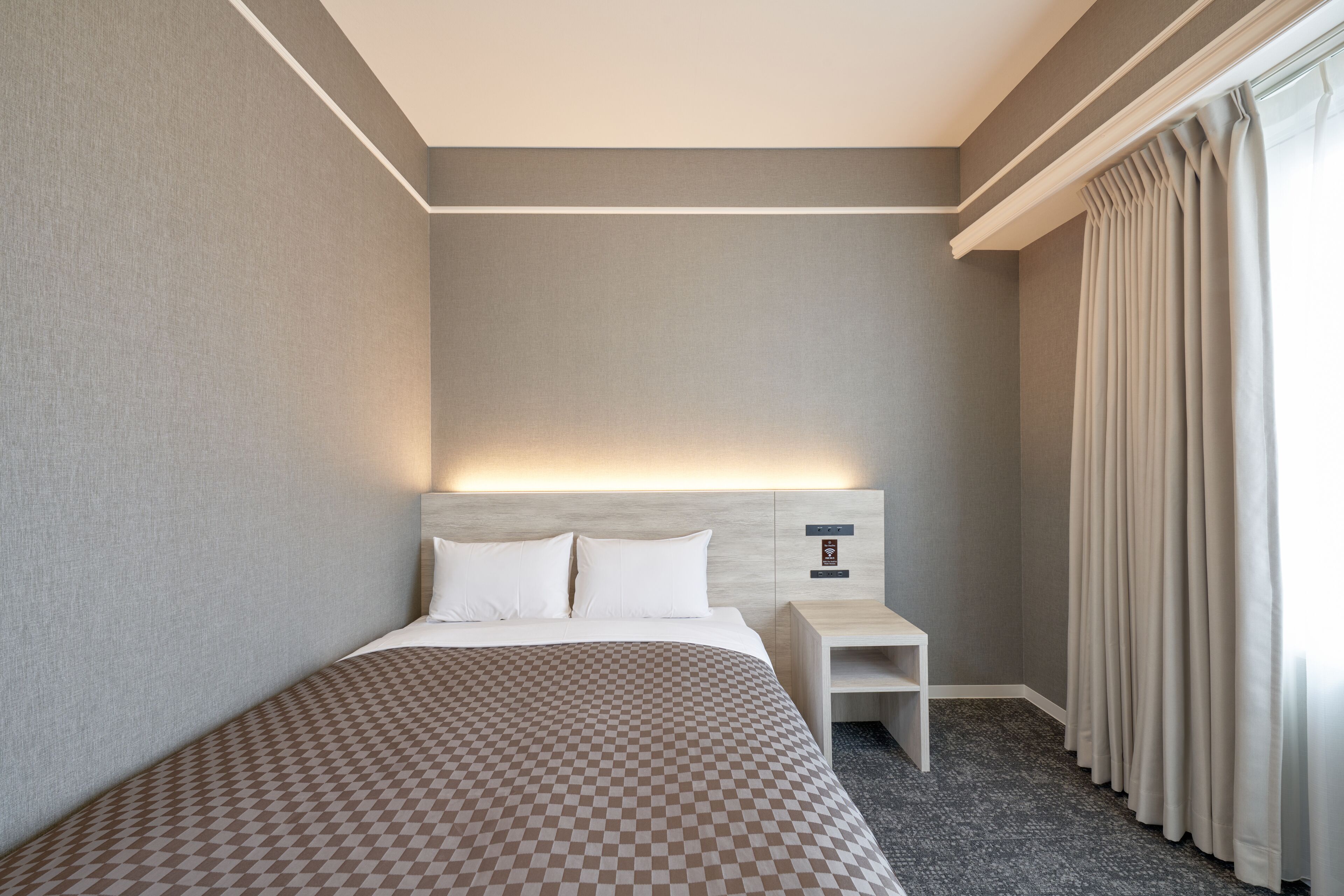 Ett hotellrum med en stor säng, ett sängbord, en liten avlastningsyta, en vägglampa och ett fönster med gardiner.
