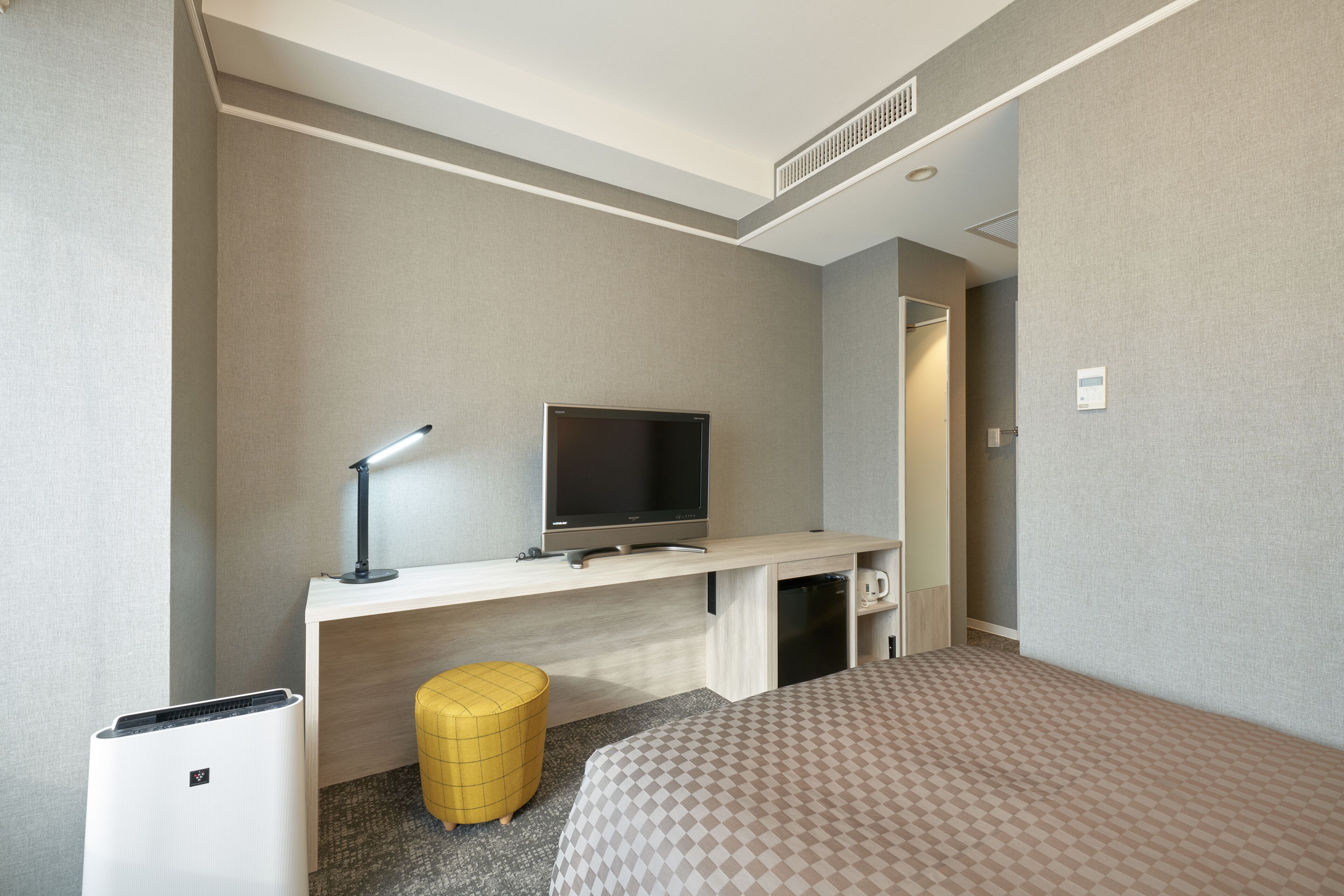 A hotel room with a bed, a desk with a television, a lamp, a small stool, and a mini-fridge.