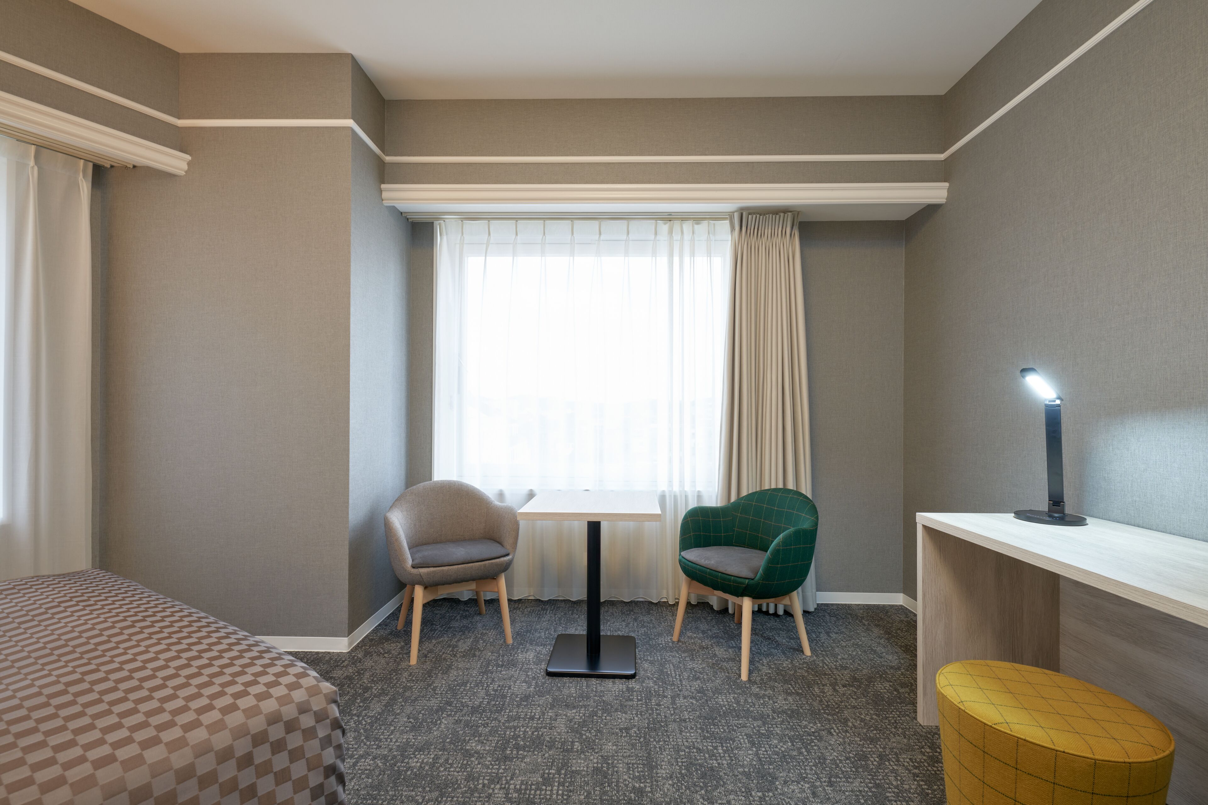 Een hotelkamer met een bed, twee stoelen, een klein tafeltje, een bureau, een lamp en een raam met gordijnen.