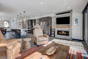 A modern living room with a fireplace, a dining area with chairs, and a kitchen with dark cabinets and a stone accent wall. - Downtown WP River Front Villa 232 I Private Hot Tub I Great Views I Arcade I Discounted Attractions (Winter Park)