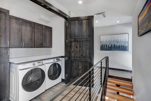 Un lavadero con dos lavadoras Samsung, una puerta de madera y una escalera con barandilla de metal.