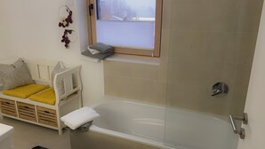 A bathroom with a bathtub, a window with a view, and a white chair with cushions. - Apartment and studio Weißensee (Füssen)