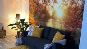 A modern living room with a dark grey sofa, yellow cushions, a large yellow rug, and a grey armchair. A large painting of autumn leaves is on the wall, and a potted plant is on the floor. - Apartment and studio Weißensee (Füssen)