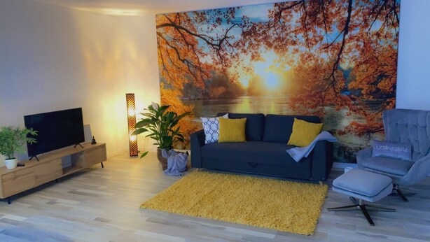 A living room with a large painting of autumn leaves, a dark blue sofa with yellow cushions, a grey armchair, a wooden TV stand, and a yellow rug. - Apartment and studio Weißensee (Füssen)