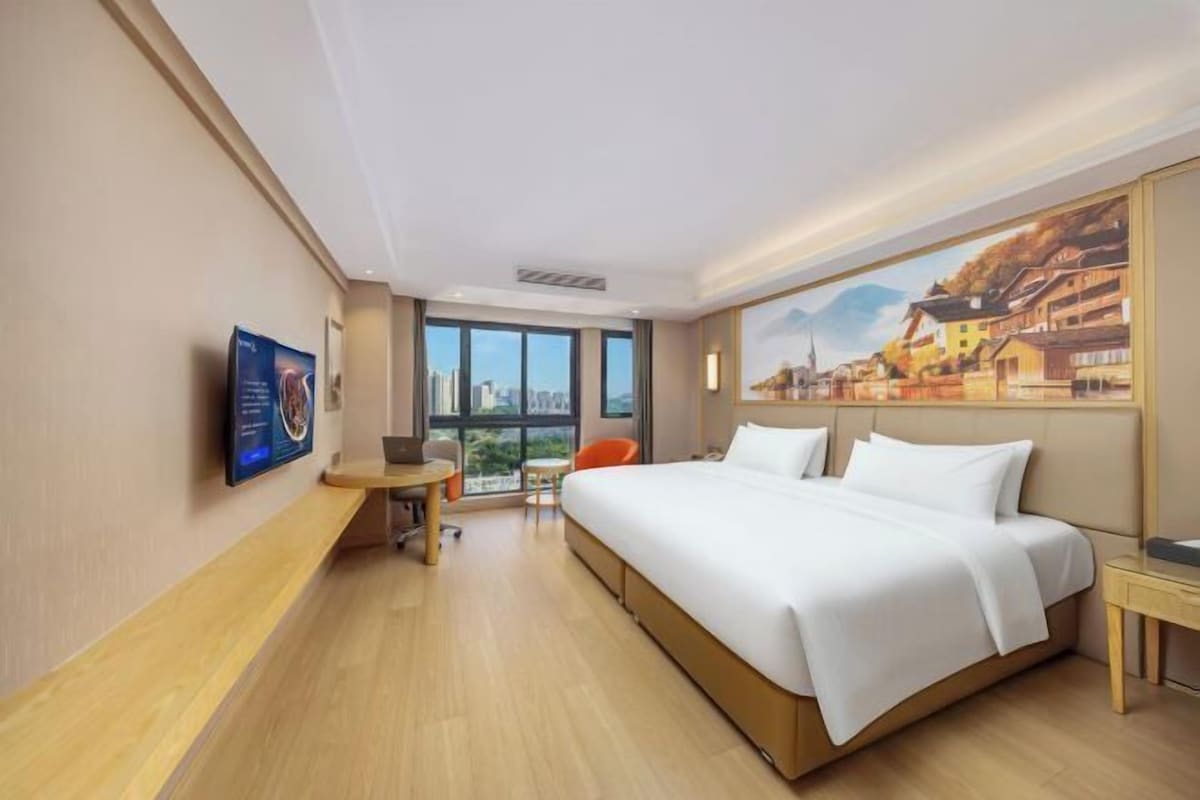 A hotel room with a large bed, a desk, a chair, a TV, and a window with a city view.
