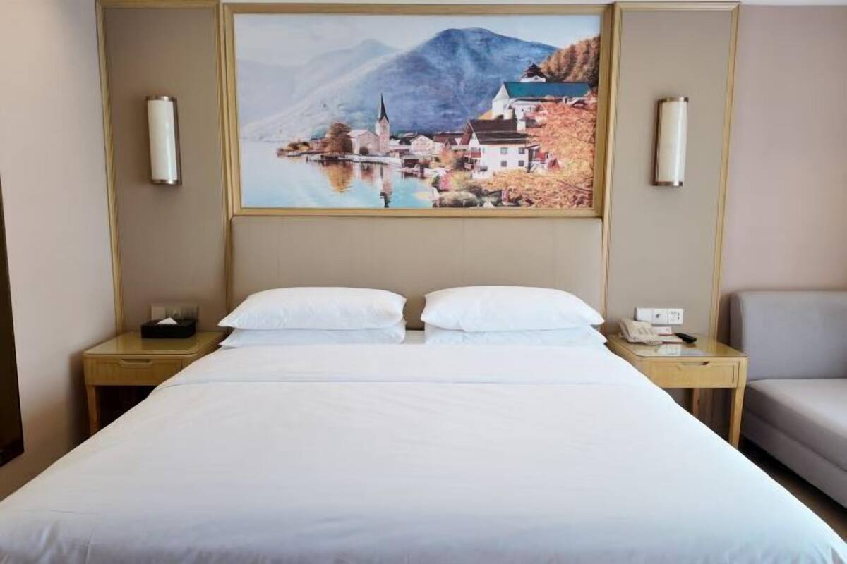 Ein Hotelzimmer mit einem großen Bett, zwei Nachttischen, einem Sessel und einem Landschaftsbild.